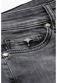 Tmavě šedé džínové kalhoty mají přední kapsu s dekorativním stříbrným prvkem, viditelné švy a hladkou texturu.