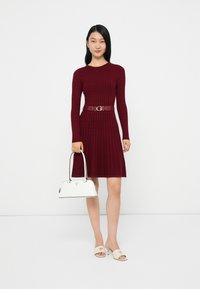 Abito a maglia bordeaux a coste con maniche lunghe, stretto in vita da una cintura coordinata. La modella tiene in mano una borsa bianca e indossa sandali beige.
