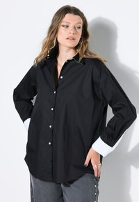 Donna che indossa una camicia nera con bottoni e dettagli bianchi sui polsini e sul colletto, pantaloni grigi con bottoni laterali, posando di fronte a uno sfondo bianco.