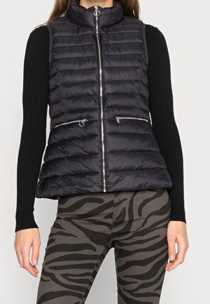 Zwarte doorgestikte mouwloze vest met ritssluiting aan de voorkant en met rits gesloten zakken, gedragen over een zwart geribd shirt met lange mouwen, gecombineerd met grijze broek met zebraprint.