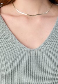 Femme portant un pull en tricot vert pâle à col en V et un collier fin, lisse et argenté autour du cou.