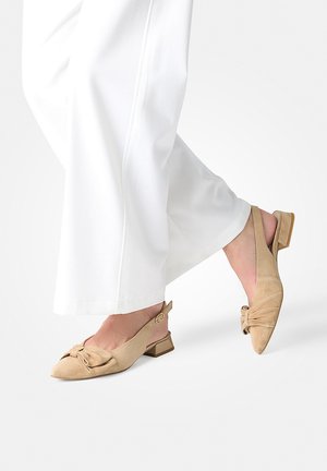 Footwear featuring beige pointed slingback flats with a knot detail, worn with white wide-leg pants against a plain background.