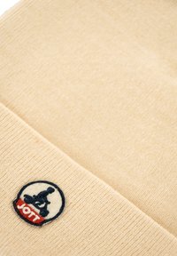 Tessuto a maglia beige con una piccola toppa ricamata che presenta un design in bianco e nero e testo rosso in basso.