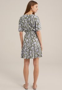 Rochie florală verde cu mâneci scurte, tiv cu volane în straturi și talie ajustată. Prezintă un model cu flori albe și purpurii. Sandale plate.