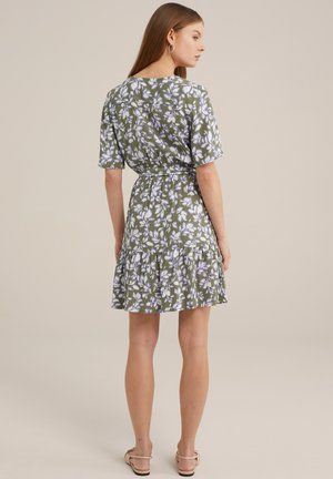 Grünes Blumenkleid mit kurzen Ärmeln, gestuftem Rüschensaum und taillierter Taille. Es zeigt ein Muster aus weißen und lila Blumen. Flache Sandalen.