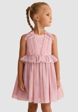 Ragazza giovane con capelli treccia indossando un vestito rosa senza maniche con volant e decorazioni di perle, che guarda verso destra su uno sfondo semplice.