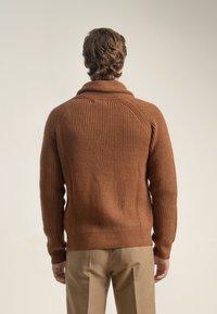 Suéter de punto acanalado marrón con cuello chal, que presenta un acabado texturizado y mangas ajustadas, utilizado con pantalones de color claro.