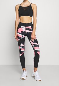 Sort sportsbh med mesh-paneler, parret med tætsiddende leggings i pink, sort og hvide geometriske mønstre. Hvide træningssko.