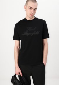 Juodas trumpas rankovėmis marškinėlis su toniniais užrašais „Karl Lagerfeld“ priekyje, pagamintas iš minkštos, lygios medžiagos. Modelis laiko mažą rankinę.