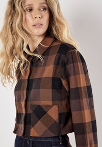 Brown and black plaid cropped jacket with a collar, zipper, and front pocket. Made of fabric with a textured finish.