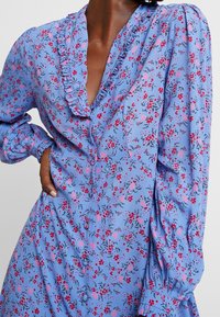 Světle modré látkové šaty s květinovým vzorem v růžové a černé barvě, s volánkovým límcem, zapínáním na knoflíky a dlouhými nařasenými rukávy.