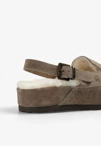 Zapato de ante marrón con plataforma plana, correa ajustable y suave forro de piel de oveja. Presenta remaches decorativos a lo largo del costado.