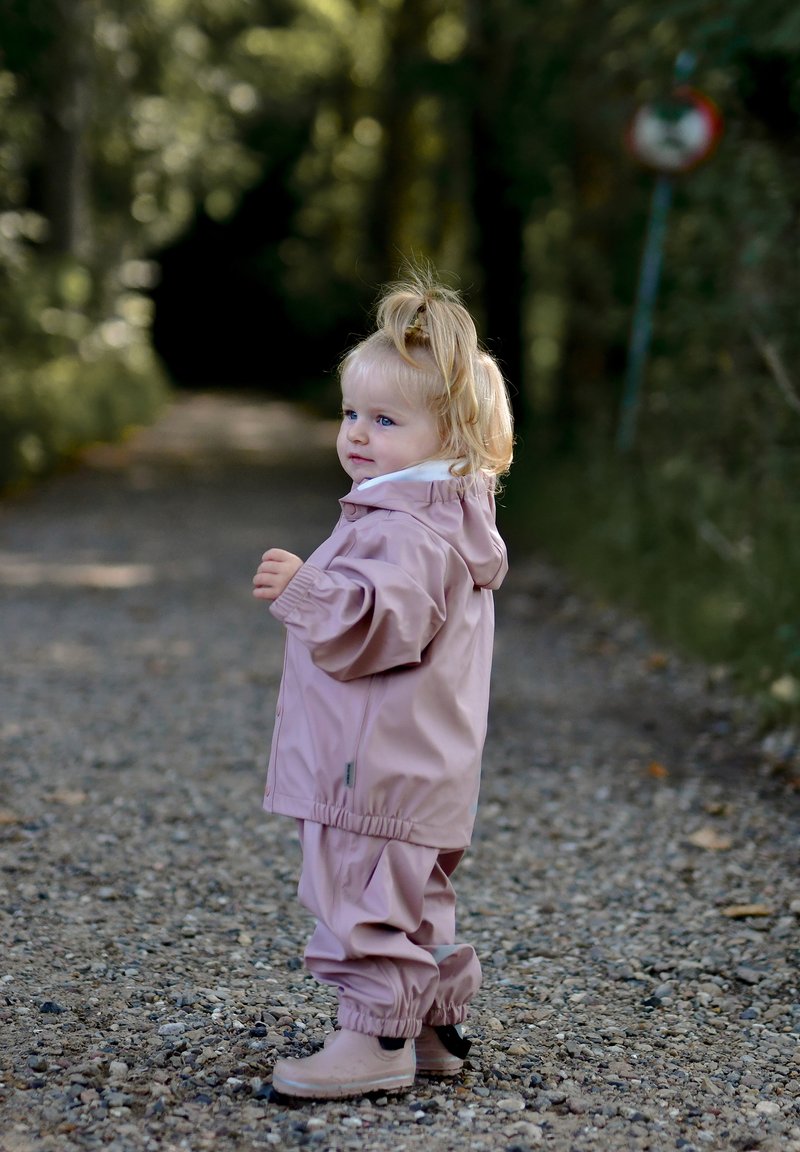 Rožinė vandeniui atspari striukė ir derančios kelnės iš lygios medžiagos; minkšta tekstūra; vaikas stovi ant žvyro tako, fone matyti medžiai.