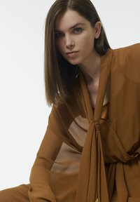 Haut en tissu transparent marron avec un accent noué au niveau du col, présentant une texture lisse et un design drapé, porté par-dessus une couche de fond marron uni.