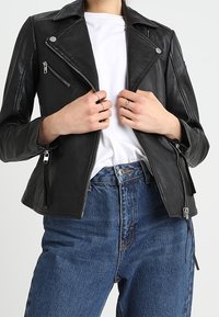 Black leather motorcycle jacket with silver hardware, buttoned lapels, and zippered pockets, paired with a white shirt and blue jeans.