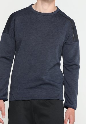 Mann trägt ein dunkelgraues Langarm-Sweatshirt mit Rundhalsausschnitt und schwarze Sporthose, steht vor einem einfarbigen hellen Hintergrund.
