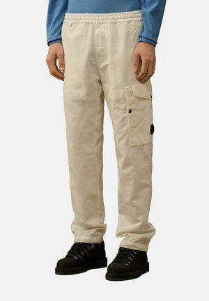 Pantalons cargo couleur crème avec une taille élastique, poche latérale avec rabat et accents en métal noir. Portés avec des bottes noires à lacets.