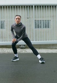 Žena u sportskoj odjeći isteže noge široko na popločanom tlu pokraj metalne ograde i betonskog zida na otvorenom.