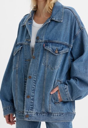 Frau trägt übergroße blaue Jeansjacke mit Knopfverschluss und Klappentaschen, kombiniert mit hellblauen Jeans und weißem Hemd.