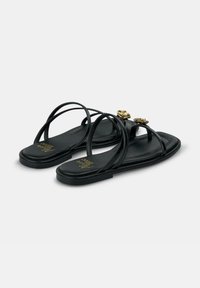 Las sandalias planas negras cuentan con correas delgadas, dos acentos en forma de rosa dorados y un acabado mate suave sobre una suela acolchada.