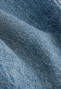 Tejido de mezclilla en azul medio con un tejido texturizado, que presenta costuras visibles y un ligero brillo. Suave y maleable con una apariencia uniforme.