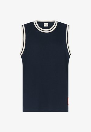 Marineblaues ärmelloses Oberteil mit weißem Besatz, kombiniert mit mehrfarbigen Badeshorts mit geometrischem Muster in Rot, Orange und Beige. Schwarze Flip-Flops.