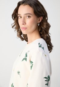 Lysfarget hettegenser med grønn blomstermønster, rund halslinning og myk tekstur. Krøllete hår og øreringer komplementerer designet.