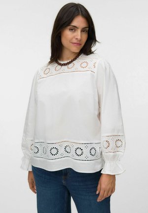 Kvinde med mørkt hår iført en hvid langærmet bluse med cirkulære blonde mønstre og blå jeans mod en ensfarvet baggrund.