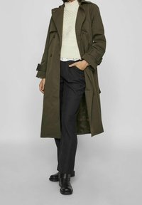 Oliwkowy płaszcz trench z podwójnym zapięciem, z paskiem w talii i naramiennikami, noszony na kremowym swetrze dzianinowym i czarnych spodniach.