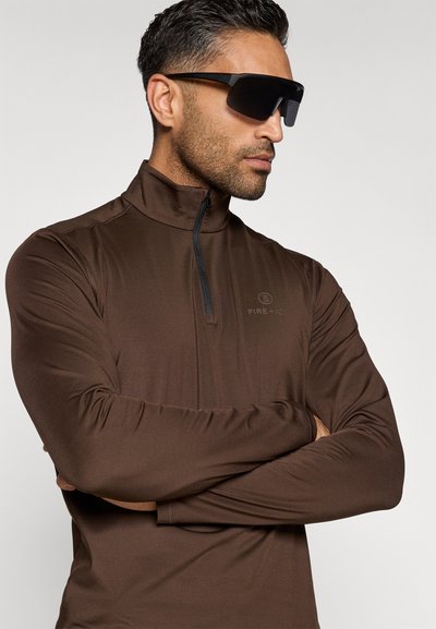 Man som bär en mörkbrun långärmad zip-up tröja med "FIRE + ICE" logotyp och svarta wraparound solglasögon, med armarna i kors, vänd mot höger mot en vit bakgrund.