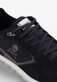 Pantof cu toc negru, realizat dintr-un amestec de piele întoarsă și materiale textile, cu un grafic de craniu gri, șireturi texturate și o talpă albă.