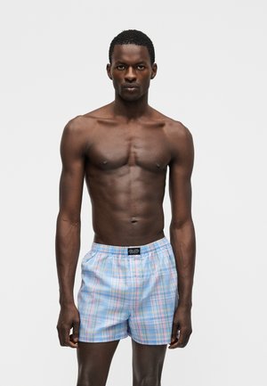 Muskulózny muž stojaci čelom, oblečený v modrých a ružových károvaných boxerkách na jednoduchom bielom pozadí.