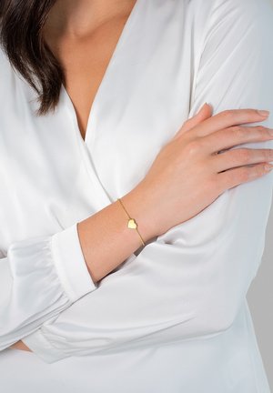 Bracelet en or avec un charm en forme de cœur, arborant un design de chaîne délicat. Porté sur un poignet au-dessus d'un haut à manches longues blanc. Texture lisse.