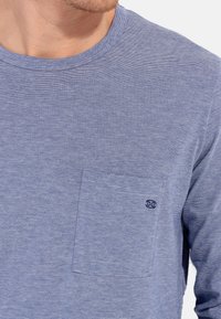 Langærmet, lyseblå t-shirt med en subtil stribet tekstur, der har en lomme på brystet og et lille logo på lommen.