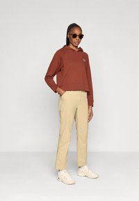 Brun cropped hettegenser med hette og logopatch, beige bukser med sidelommer og beige sneakers; modellen har på seg mørke solbriller.