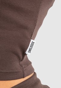 Brun ribbad tyg med sid sömmar och en vit etikett märkt "SMILODOX" fäst. Designen skapar en figurnära silhuett.