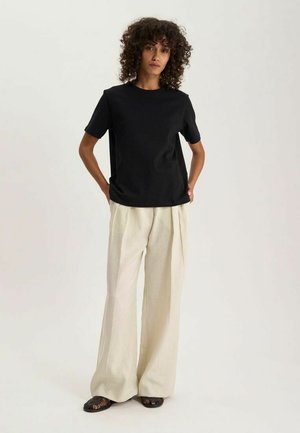 Femme aux cheveux bouclés portant un t-shirt noir à manches courtes, un pantalon beige ample et des chaussures sombres tissées, debout les mains dans les poches.