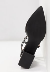 Escarpin noir en cuir verni à talon bloc et bout pointu, semelle tournée vers l’avant sur fond blanc.