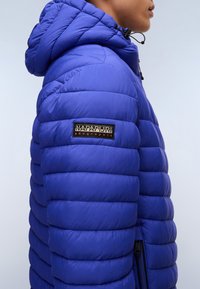 Modrá lehká bunda s kapucí, strukturovaným prošívaným designem a výrazným logem našitým na rukávu. Detail s zipem na boku.