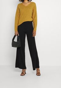 Musztardowy sweter z dekoltem w kształcie litery V, połączony z czarnymi szerokimi spodniami w prążki. Zawiera małą plecioną czarną torebkę oraz czarne sandały na paski.