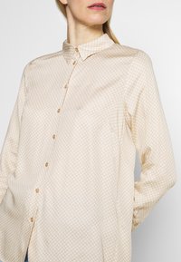 Pale beige button-up shirt with a regular collar, featuring a subtle white polka dot pattern, long sleeves, and a relaxed fit.