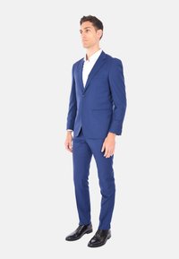 Completo tailored blu navy con taglio aderente, giacca a un bottone, pantaloni slim e scarpe nere lucide; texture del tessuto liscia.