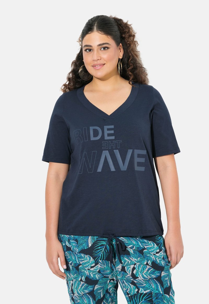 Camiseta navy de cuello en V con texto impreso "RIDE THE WAVE". Tejido suave con un corte relajado, combinada con pantalones de estampado de hojas verdes.