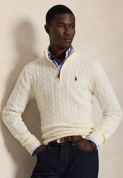 Giovane uomo con maglione bianco a trecce con mezza zip, camicia blu e jeans scuri con le mani in tasca, su uno sfondo semplice.