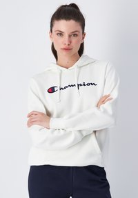 Sudadera blanca de algodón con un bolsillo tipo canguro en el frente, capucha ajustable con cordón y logo bordado de "Champion" en azul marino y rojo.