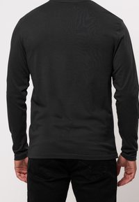 Ein Mann trägt ein schlichtes schwarzes Langarmshirt und schwarze Hosen, von hinten vor einem weißen Hintergrund gezeigt.