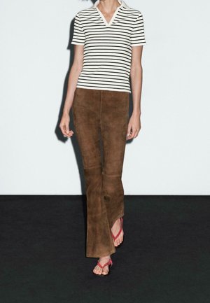 Osoba ubrana w biało-czarną koszulkę w paski na krótkie rękawy, brązowe, rozkloszowane spodnie zamszowe oraz czerwone sandały, idąca po ciemnej podłodze.