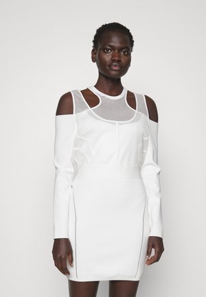 Femme aux cheveux courts et bouclés, portant une robe blanche à épaules dénudées avec des détails en mesh découpés, se tenant devant un fond blanc uni.