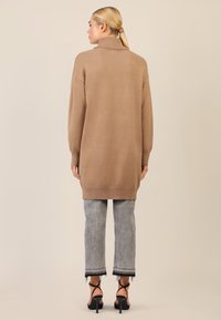 Beiger Oversized-Pullover mit hohem Kragen, gerippten Bündchen und Saum. Getragen über verwaschenen grauen Jeans und schwarzen Riemchen-Heels.