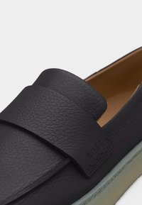 Μαύρο παπούτσι από υφή δέρματος τύπου slip-on με φαρδύ λουράκι στην κορυφή και ανοιχτό γκρι σόλα, κοντινή λήψη που δείχνει λεπτομέρειες υλικού και ραφές.